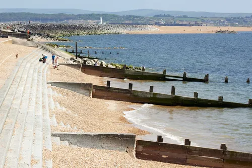 This landscape photograph captures Milford-on-sea beach in Christchurch, Dorset, United Kingdom, during the early afternoon in late spring. The image prominently features the shingle beach bordered by concrete steps and wooden groynes extending into the water. In the background, Hurst Point Lighthouse stands clearly visible, serving as a notable maritime landmark, alongside Hurst Castle which can be seen further along the coastline. The calm blue waters and distant green hills, together with beachgoers enjoying the warm weather, indicate typical late spring conditions in this scenic coastal region. The photograph highlights the relationship between the natural landscape of Milford-on-sea and the historic features of the Hurst Point Lighthouse and Castle.
